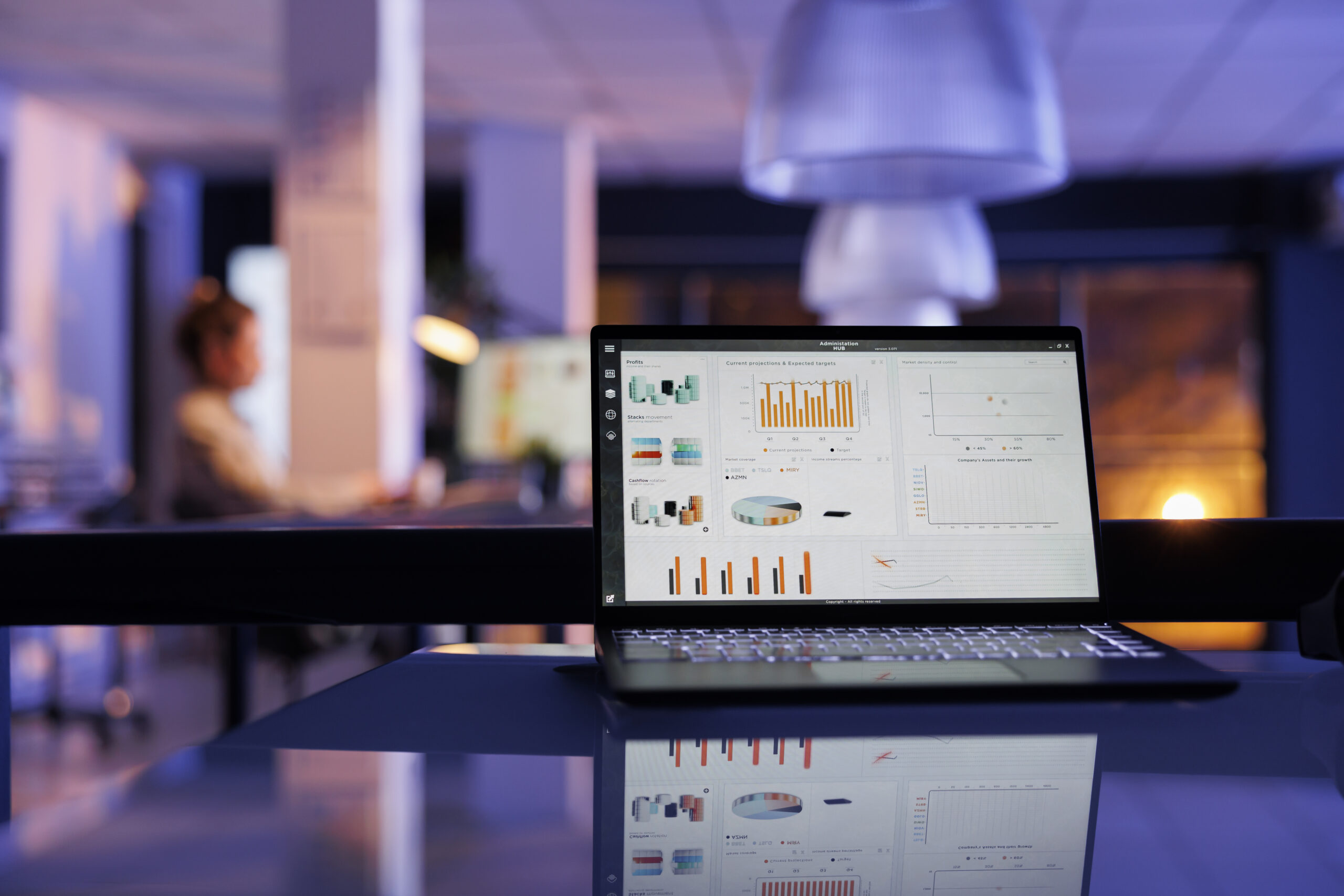 Laptop computer with company profit progress on screen standing on table in startup office. In background, businesswoman working overtime at financial strategy, analyzing marketing statistics